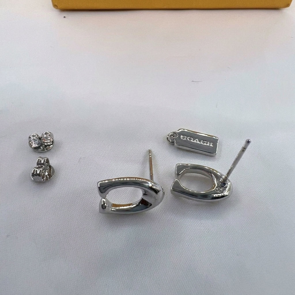 A pair of silver-tone Coach D-ring earrings shown disassembled on a white surface, with the small matching stud backs and a small rectangular box engraved with the Coach logo.