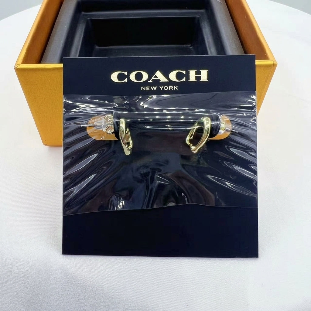 A pair of gold-tone hoop earrings displayed on a black Coach New York branded card, with the gold and black Coach box partially visible behind it on a white surface.