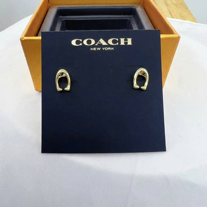 A pair of gold-tone Coach New York hoop earrings displayed on a black branded card, with an open gold box behind it on a white surface.