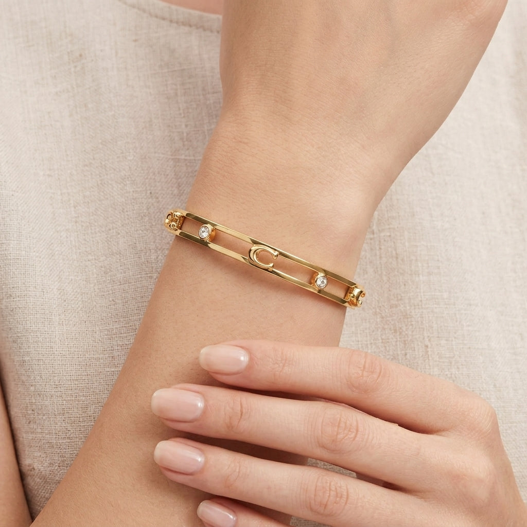 A gold-toned safety-pin style bangle bracelet with small clear crystal accents, shown worn on a woman's wrist with her hand resting below.