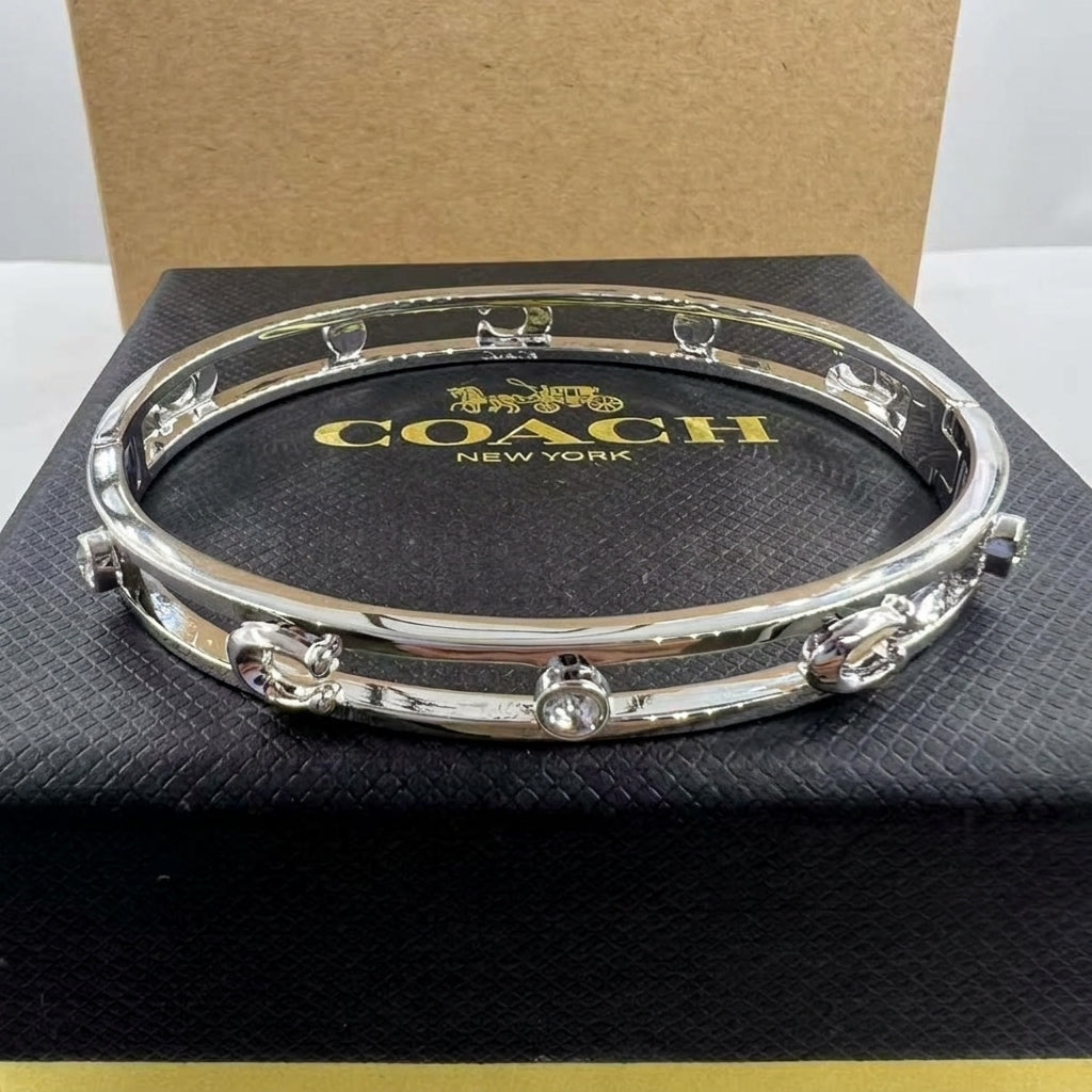A silver-tone Coach bangle bracelet with clear crystal embellishments, displayed on its black textured box with gold Coach branding.