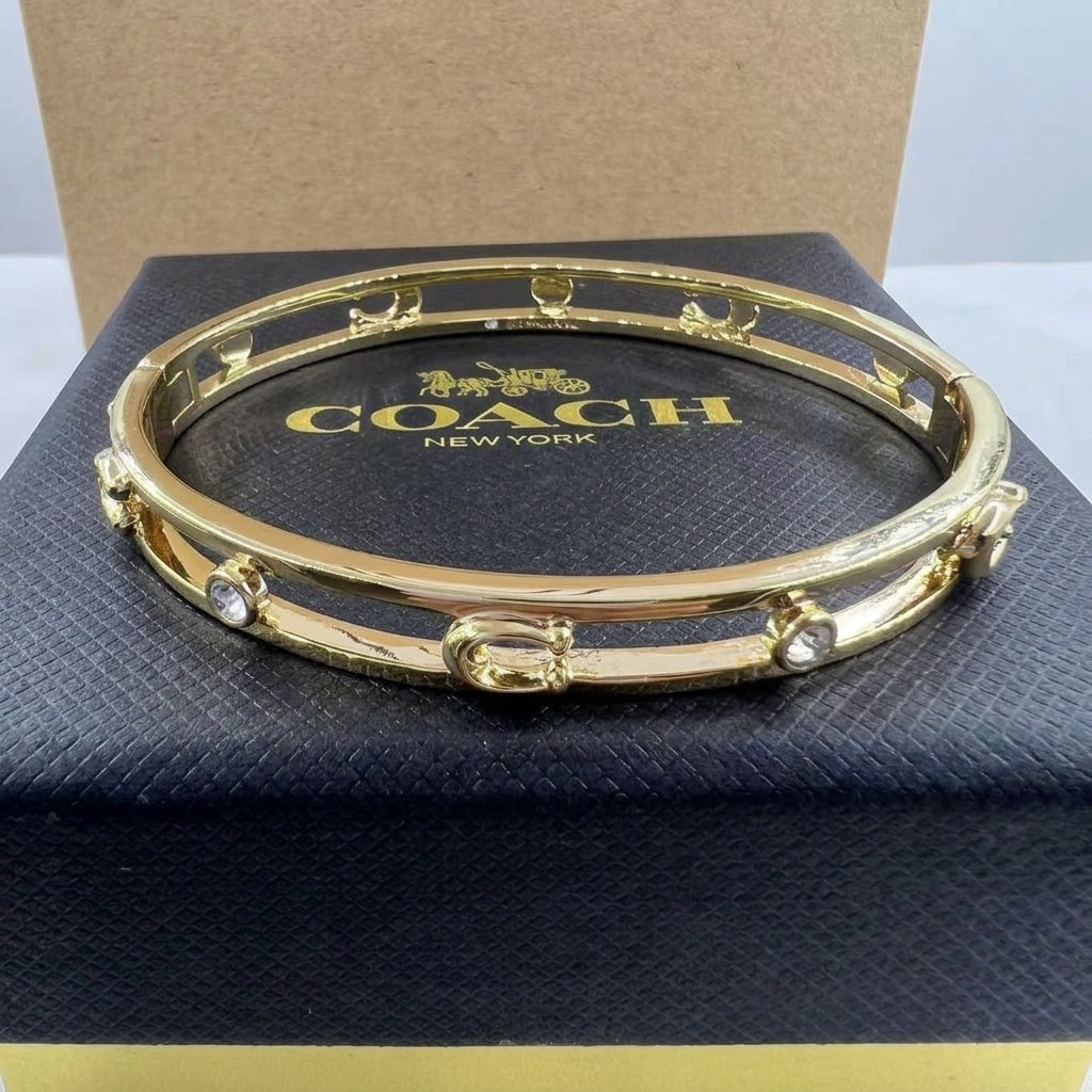 A gold-tone Coach bangle bracelet with small clear rhinestone accents, displayed on top of a black Coach New York jewelry box.