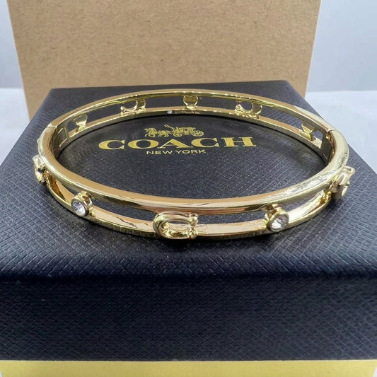 A gold-tone Coach bangle bracelet with small clear rhinestone accents, displayed on top of a black Coach New York jewelry box.
