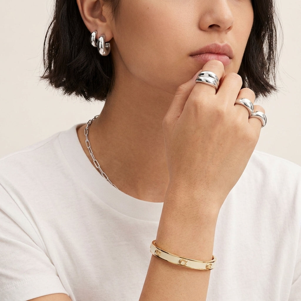 A close-up lifestyle shot of a model wearing multiple pieces of jewelry, including a gold bangle bracelet, silver hoop earrings, and silver rings, against a clean off-white background.