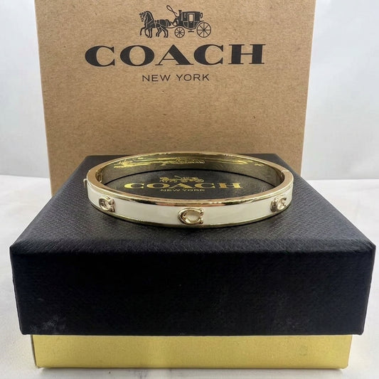 A gold and white Coach bangle bracelet displayed on a black presentation box with a brown Coach New York box behind it.