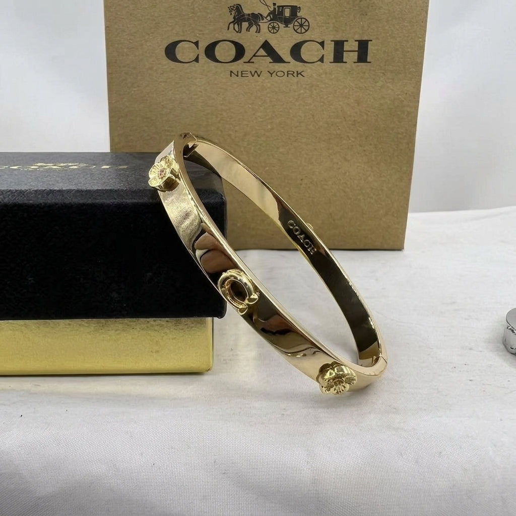 A gold-tone Coach bangle bracelet with circular floral details, displayed in front of a brown Coach New York box and a black presentation box on a white surface.