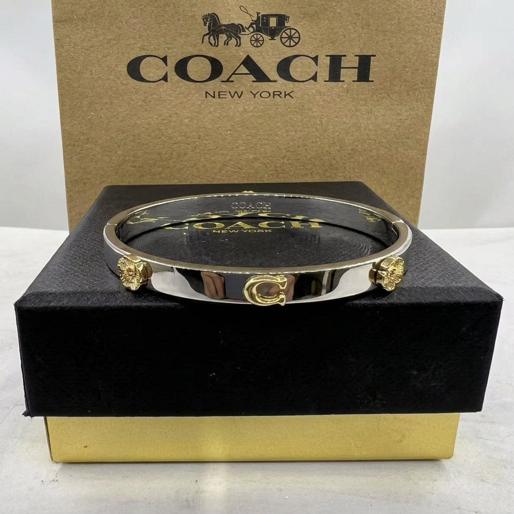 A Coach bangle bracelet with polished silver-toned metal and gold-toned lion head accents, displayed on a black box with a gold base in front of a brown Coach New York box.