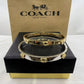 Two Coach bangle bracelets, one gold and one silver, displayed on a black presentation box with a brown Coach New York box behind them.