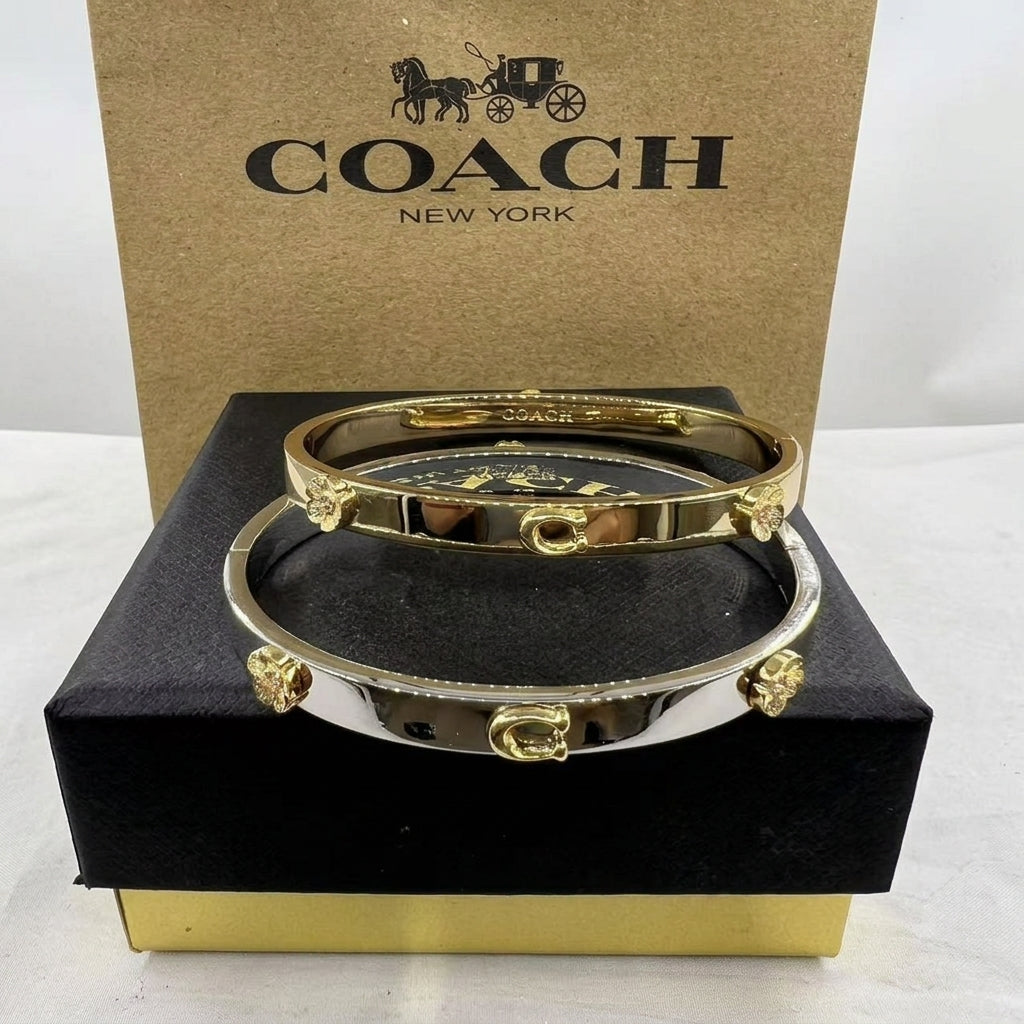 Two Coach bangle bracelets, one gold and one silver, displayed on a black presentation box with a brown Coach New York box behind them.