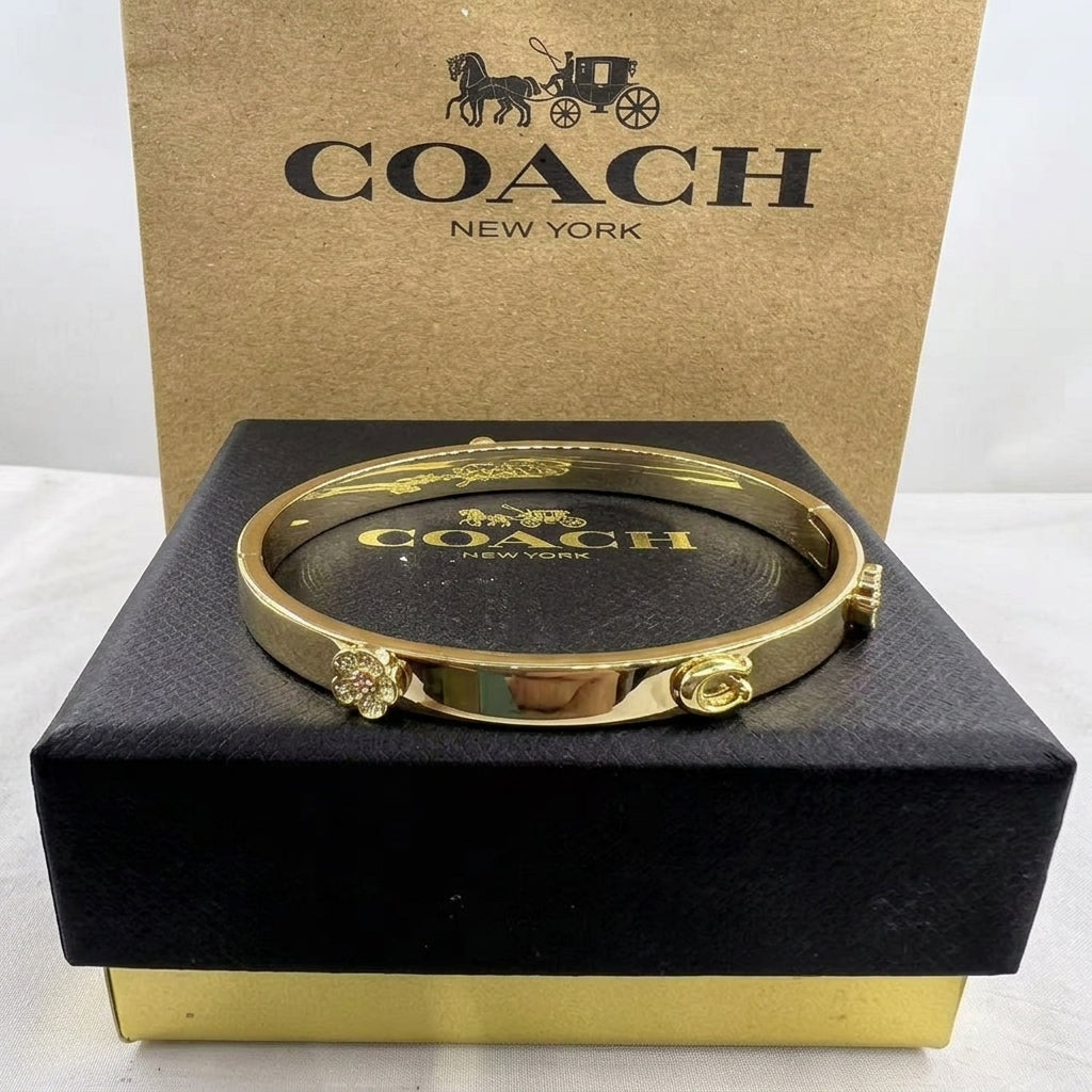 A gold-tone Coach bangle bracelet displayed on a black presentation box with a branded Coach box behind it.