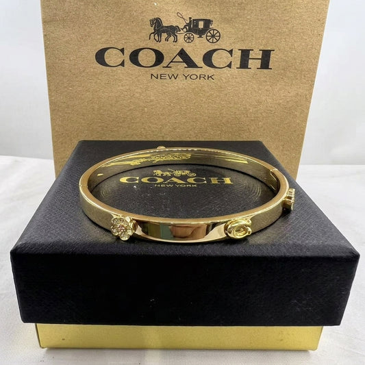 A gold-tone Coach bangle bracelet displayed on a black presentation box with a branded Coach box behind it.