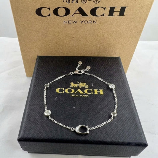 A silver-tone Coach bracelet with a horse-and-carriage charm and small clear stones, displayed on a black Coach box with a brown Coach box behind it.