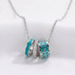 A silver chain necklace with three interlocking cylindrical charms. Two charms are turquoise blue with silver detailing, and one is silver with small clear rhinestones.