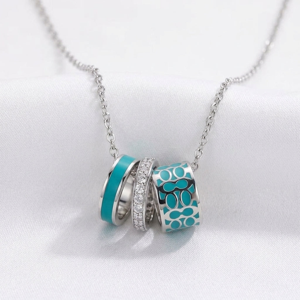 A silver chain necklace with three interlocking cylindrical charms. Two charms are turquoise blue with silver detailing, and one is silver with small clear rhinestones.