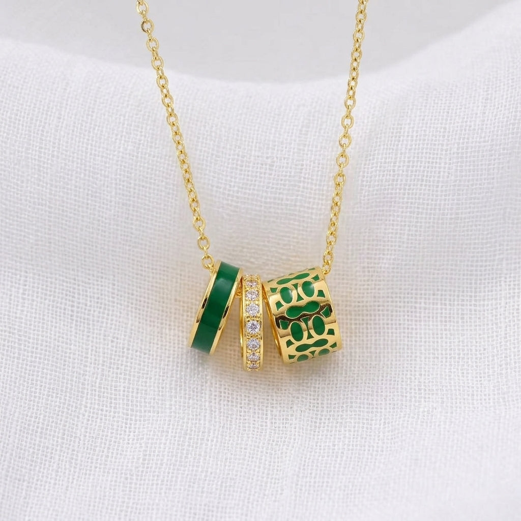 A gold chain necklace with three cylindrical charms in green and gold, displayed on a textured white fabric background.