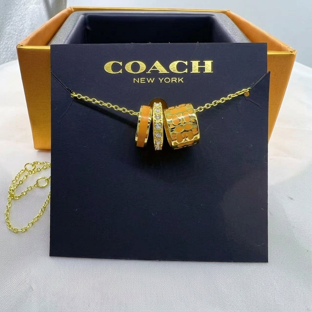 A Coach New York gold-tone charm necklace displayed on a black branded card, with the gold box behind it on a white surface.