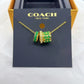 A Coach New York gold chain necklace with two cylindrical charms, one solid green and one green and gold patterned with small clear stones, displayed on a black COACH card insert.