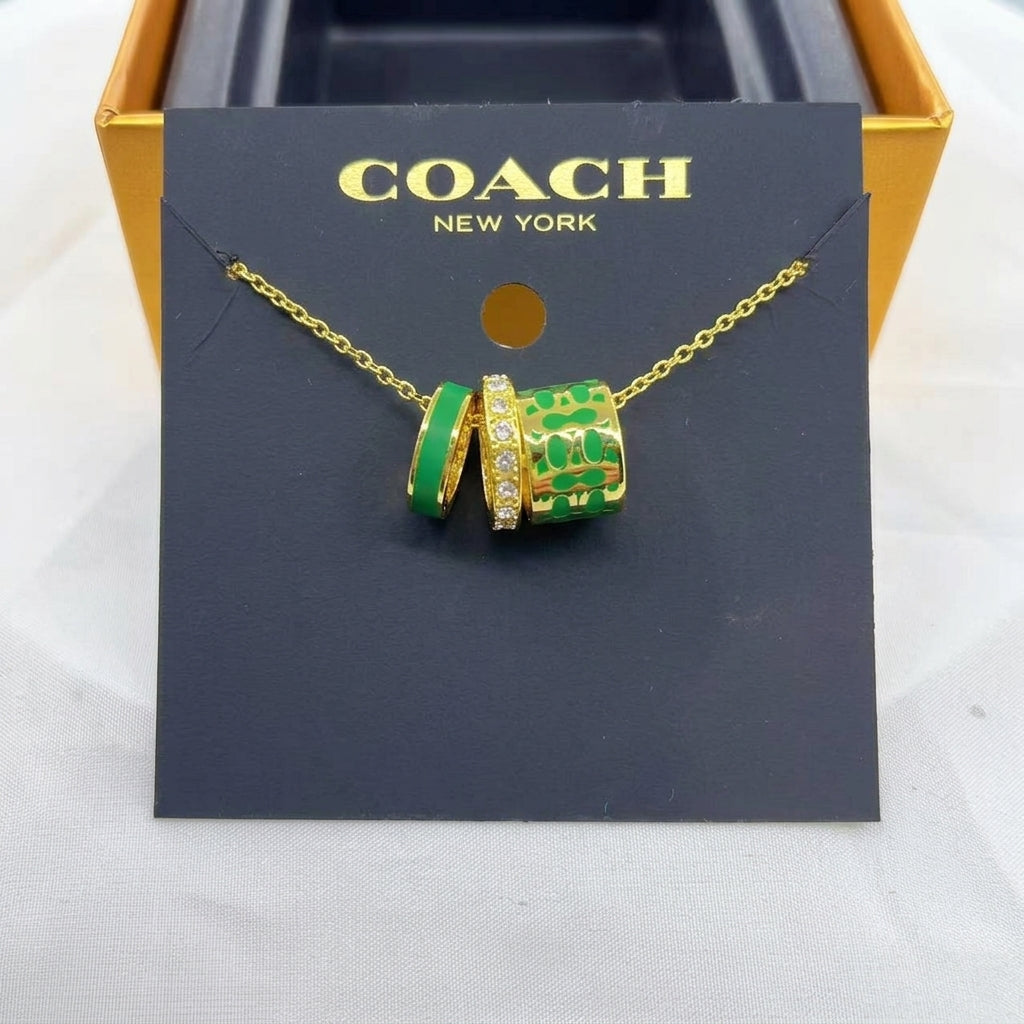 A Coach New York gold chain necklace with two cylindrical charms, one solid green and one green and gold patterned with small clear stones, displayed on a black COACH card insert.