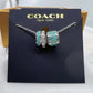 A Coach New York charm necklace displayed on a black branded card, featuring a silver-tone chain and two cylindrical charms with turquoise and black patterned details, set against a gold and black Coach box.
