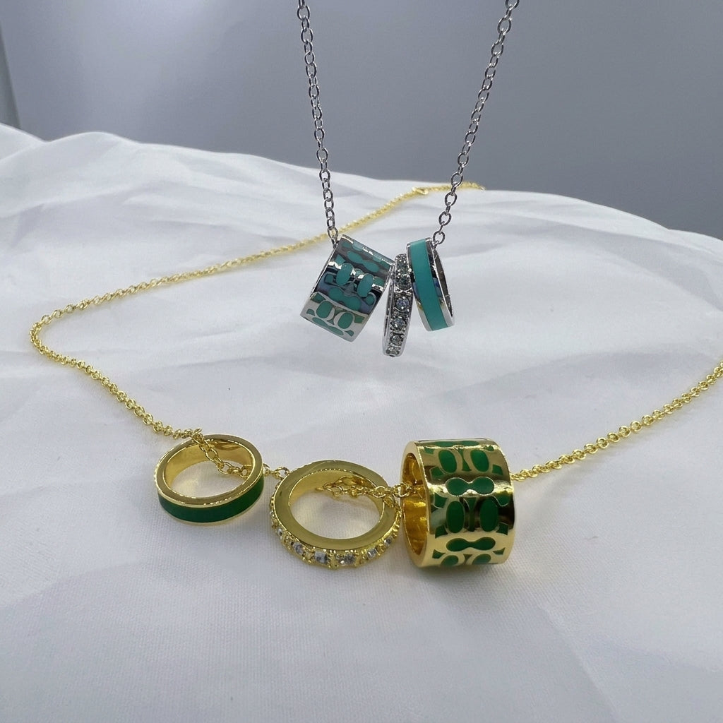 A gold chain necklace with three dangling cylindrical charms, shown draped on white fabric. The top pendant is silver with turquoise and green patterned details, while the bottom pendant is gold with a green patterned wrap and a small clear rhinestone ring charm.