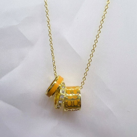 A gold chain necklace featuring a pendant with three stacked gold cylinder charms, one with bright orange-yellow enamel, and one with small clear rhinestones, displayed on a white fabric background.