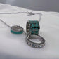 A silver-toned multi-ring charm necklace with turquoise and black enamel details, displayed on a white fabric surface.