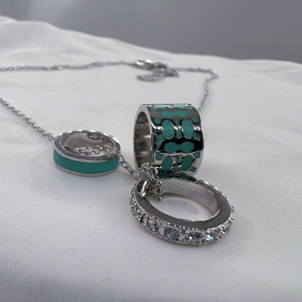 A silver-toned multi-ring charm necklace with turquoise and black enamel details, displayed on a white fabric surface.