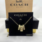 A Coach New York gold-tone necklace with three interlocking cylindrical charms, displayed on a black branded card in front of a tan Coach box.