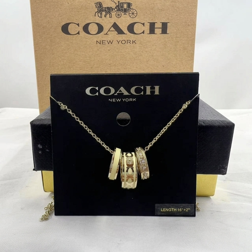 A Coach New York gold-tone necklace with three interlocking cylindrical charms, displayed on a black branded card in front of a tan Coach box.