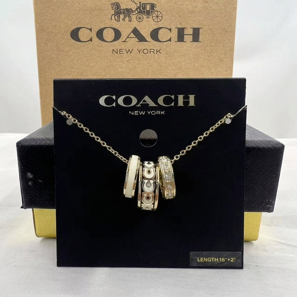 A Coach New York necklace displayed on a black branded card, with a gold-tone chain and three cylindrical charm beads, set in front of a tan Coach box and black presentation box.
