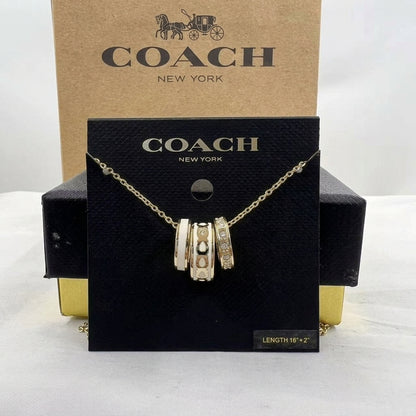 A Coach New York gold-tone charm necklace displayed on a black branded card, with the kraft paper Coach box behind it on a white studio background.