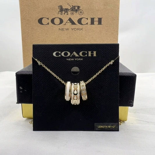 A Coach New York gold-tone charm necklace displayed on a black branded card, with the kraft paper Coach box behind it on a white studio background.