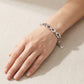 A silver link bracelet with openwork details and a small hanging tag, shown worn on a person's wrist against a light beige textured background.