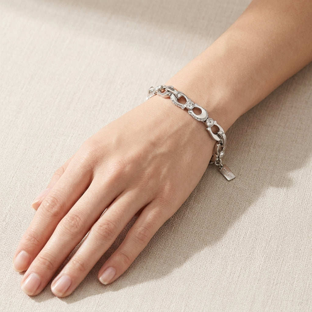 A silver link bracelet with openwork details and a small hanging tag, shown worn on a person's wrist against a light beige textured background.