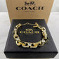 A gold-tone Coach New York bracelet with rhinestone accents, displayed on a black textured Coach New York box with its kraft paper box behind it.