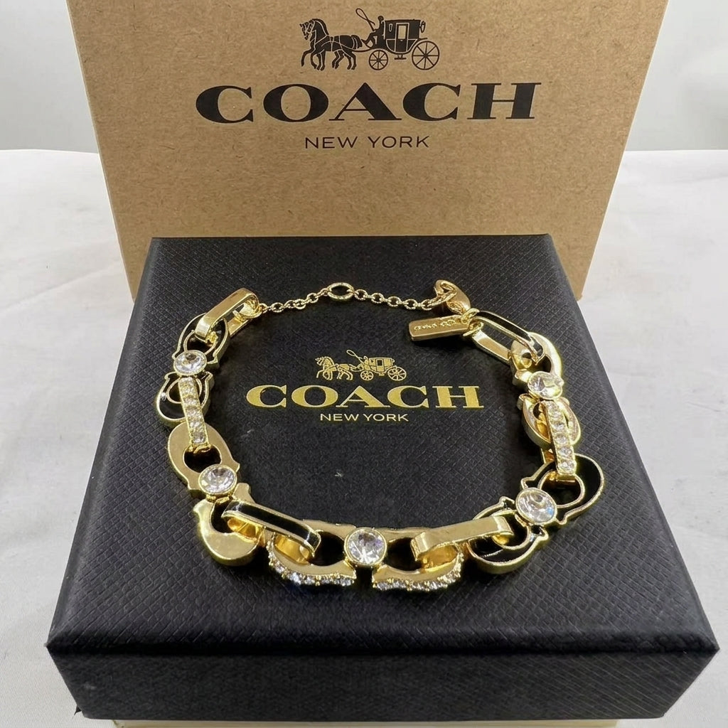 A gold-tone Coach New York bracelet with rhinestone accents, displayed on a black textured Coach New York box with its kraft paper box behind it.