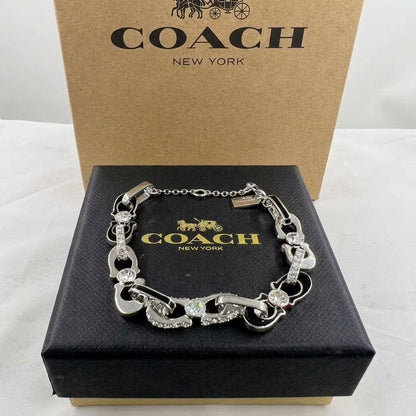 A silver-tone Coach bracelet with horsebit links and clear crystal accents, displayed on a black Coach New York presentation box with the original brown cardboard box behind it.