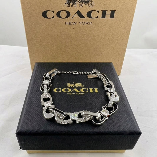 A silver-tone Coach bracelet with horsebit links and clear crystal accents, displayed on a black Coach New York presentation box with the original brown cardboard box behind it.
