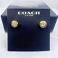 A pair of gold-tone Coach New York stud earrings with clear crystal centers, mounted on a dark navy blue branded display card in front of a matching Coach box.