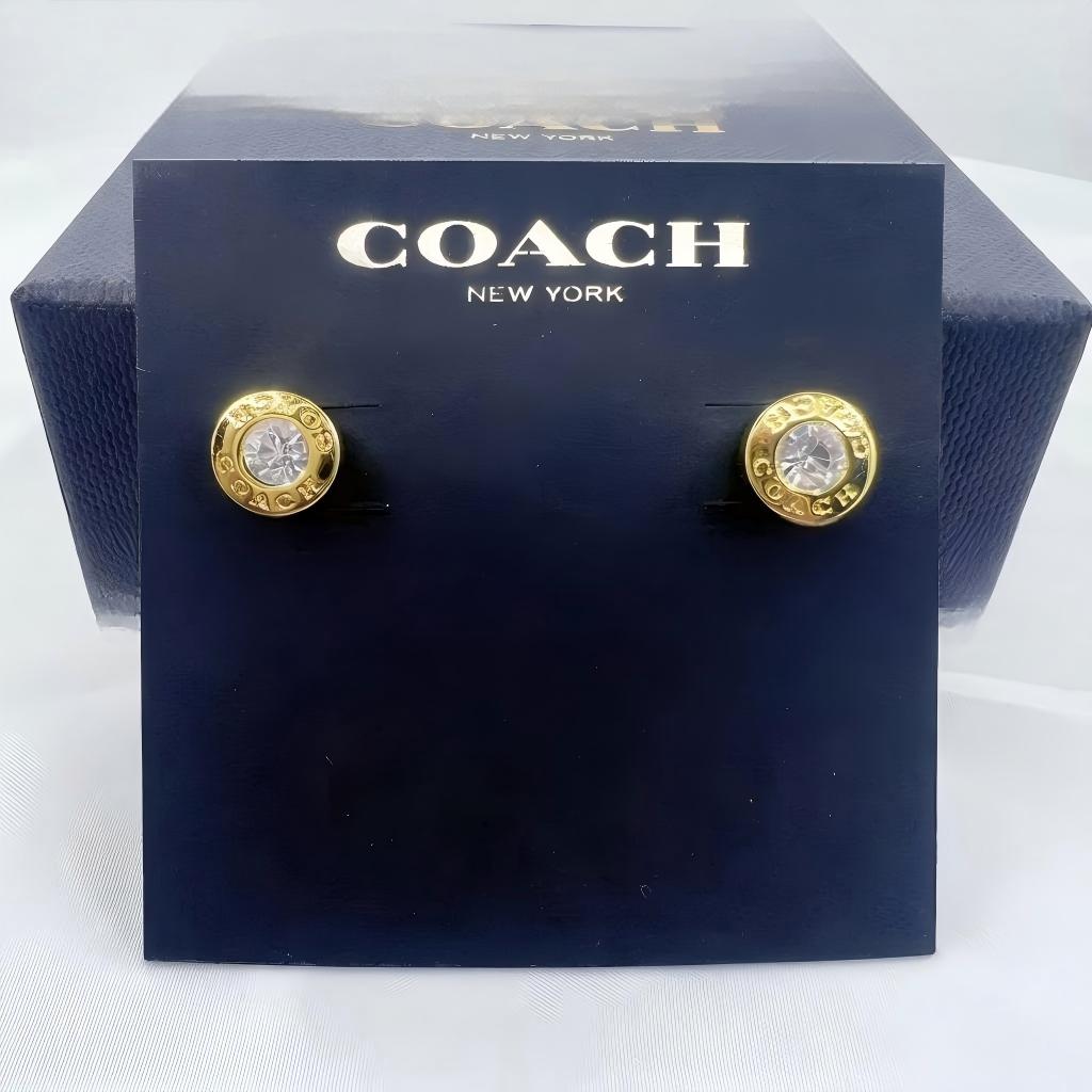 A pair of gold-tone Coach New York stud earrings with clear crystal centers, mounted on a dark navy blue branded display card in front of a matching Coach box.
