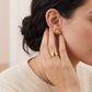 A close-up lifestyle shot of a model wearing a textured gold dome stud earring on her ear and a polished gold band ring on her finger, wearing a cream-colored knit sweater.