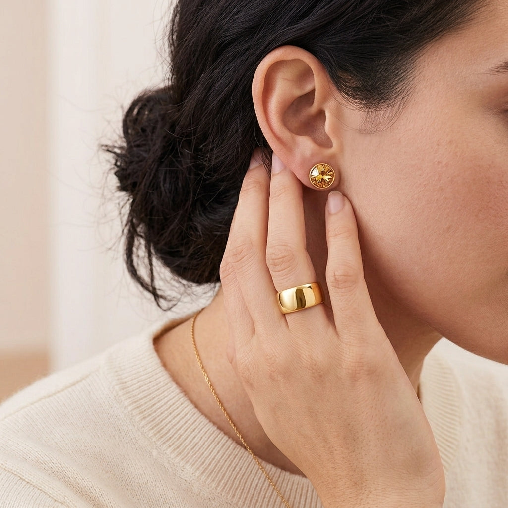 A close-up lifestyle shot of a model wearing a textured gold dome stud earring on her ear and a polished gold band ring on her finger, wearing a cream-colored knit sweater.