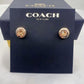 A pair of Coach New York stud earrings displayed on a branded dark navy presentation card, with the Coach box partially visible behind it.
