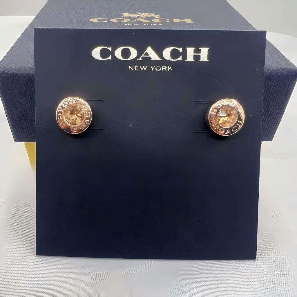 A pair of Coach New York stud earrings displayed on a branded dark navy presentation card, with the Coach box partially visible behind it.