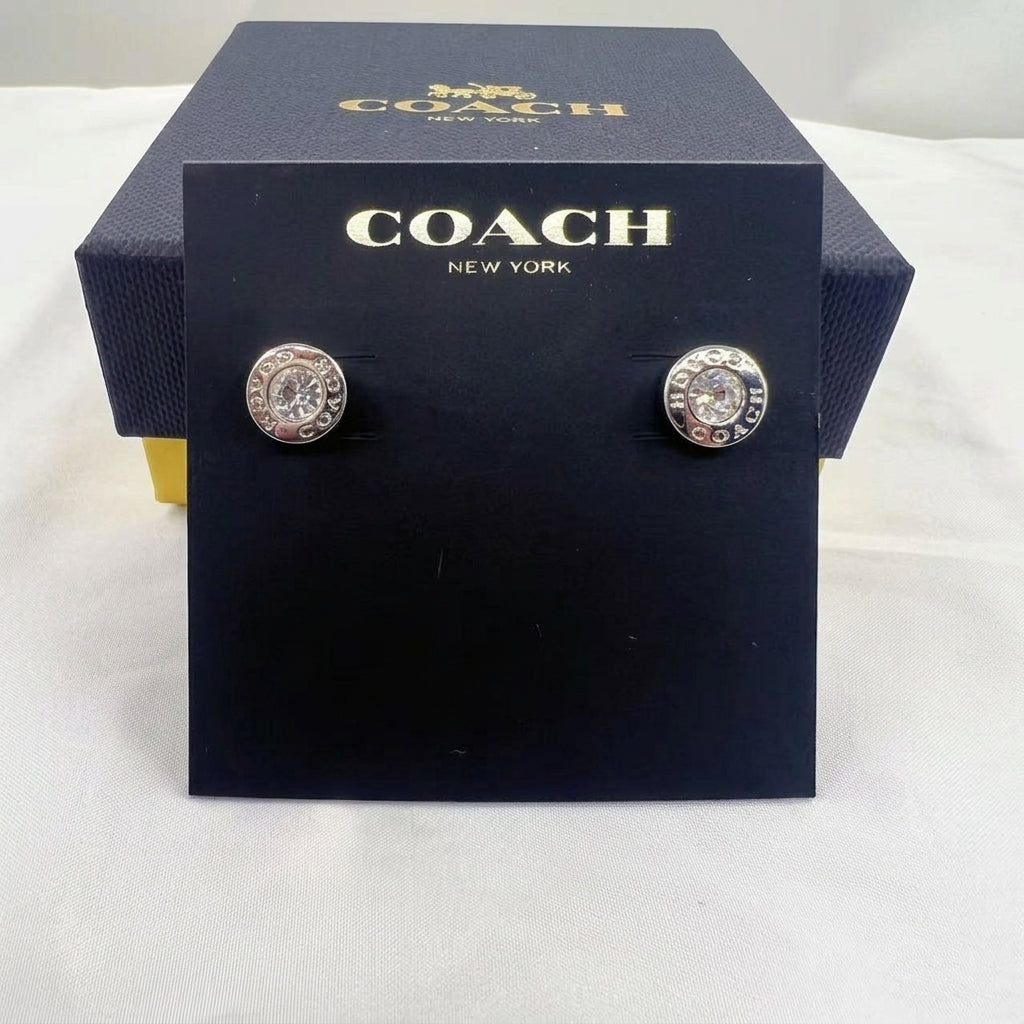 A pair of Coach New York stud earrings displayed on a black branded card, with the matching Coach box behind it on a white fabric surface.