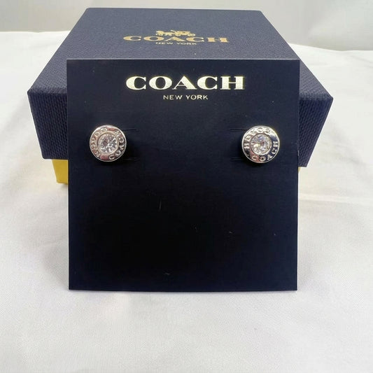 A pair of Coach New York stud earrings displayed on a black branded card, with the matching Coach box behind it on a white fabric surface.