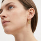 A close-up of a woman's ear wearing a small gold dangling earring with an intricate filigree pattern, set against a clean white background.