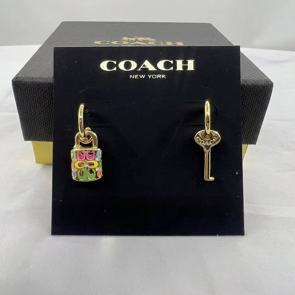 A pair of Coach New York drop earrings displayed on a black branded card, with the black Coach box behind it. One earring is a colorful lock charm with pink, green, yellow, and blue enamel details, and the other is a gold key-shaped charm.