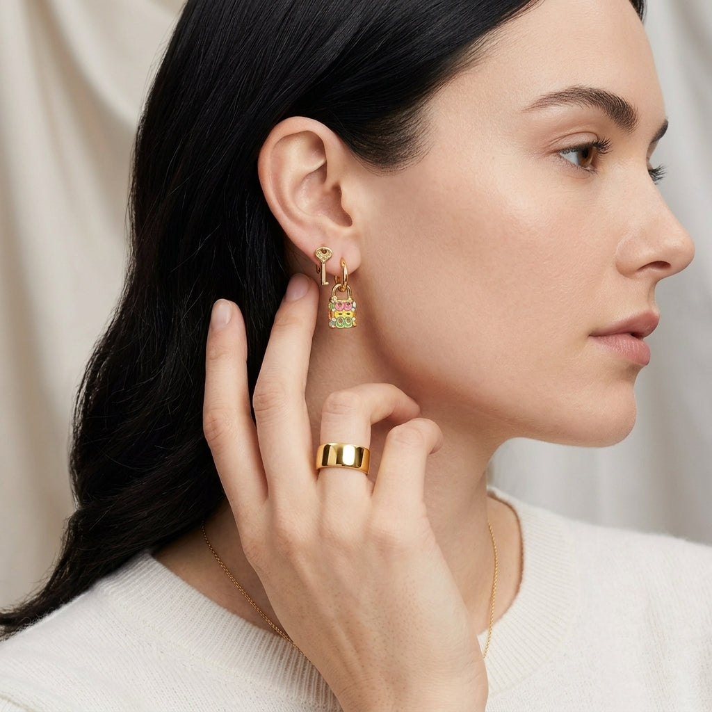 A close-up side profile of a woman with dark hair wearing a gold hoop earring with a small colorful charm and a wide gold ring, against a soft beige draped background.