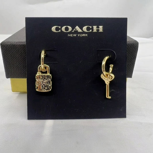 A pair of gold-tone Coach earrings displayed on a black branded card, with a Coach box partially visible behind. One earring is a padlock charm and the other is a key-shaped charm.